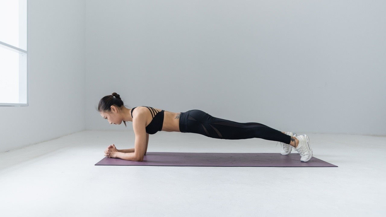 A female in black exercise clothes holding a plank on a yoga mat in a white room