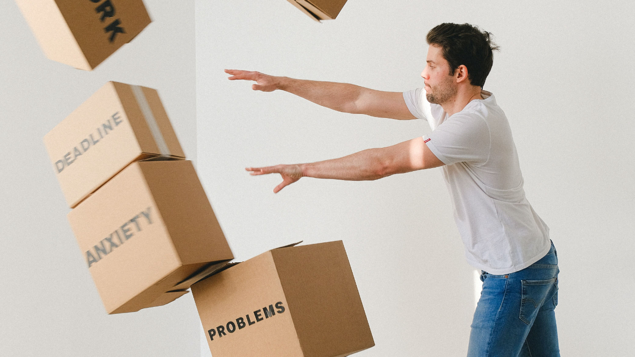 A man wearing a white t-shirt and blue jeans, pushing boxes labelled 'deadline', 'anxiety', 'problems' over.