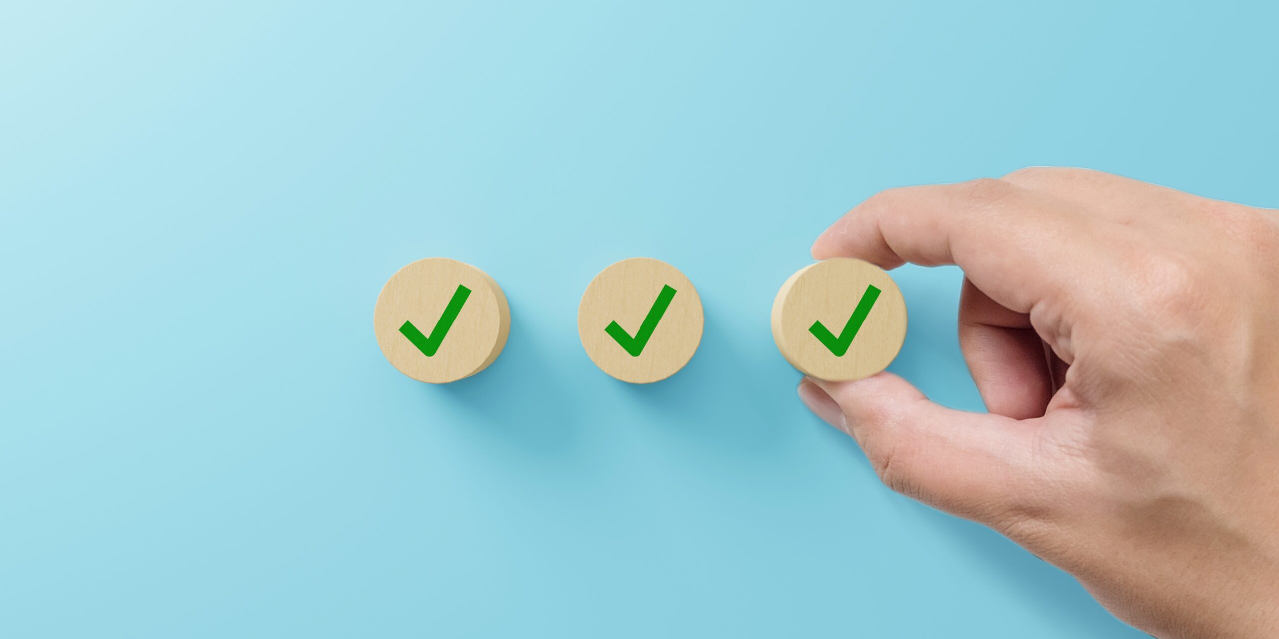 Three wooden blocks with a green tick on each of them on a blue background. Two fingers are holding the third tick.