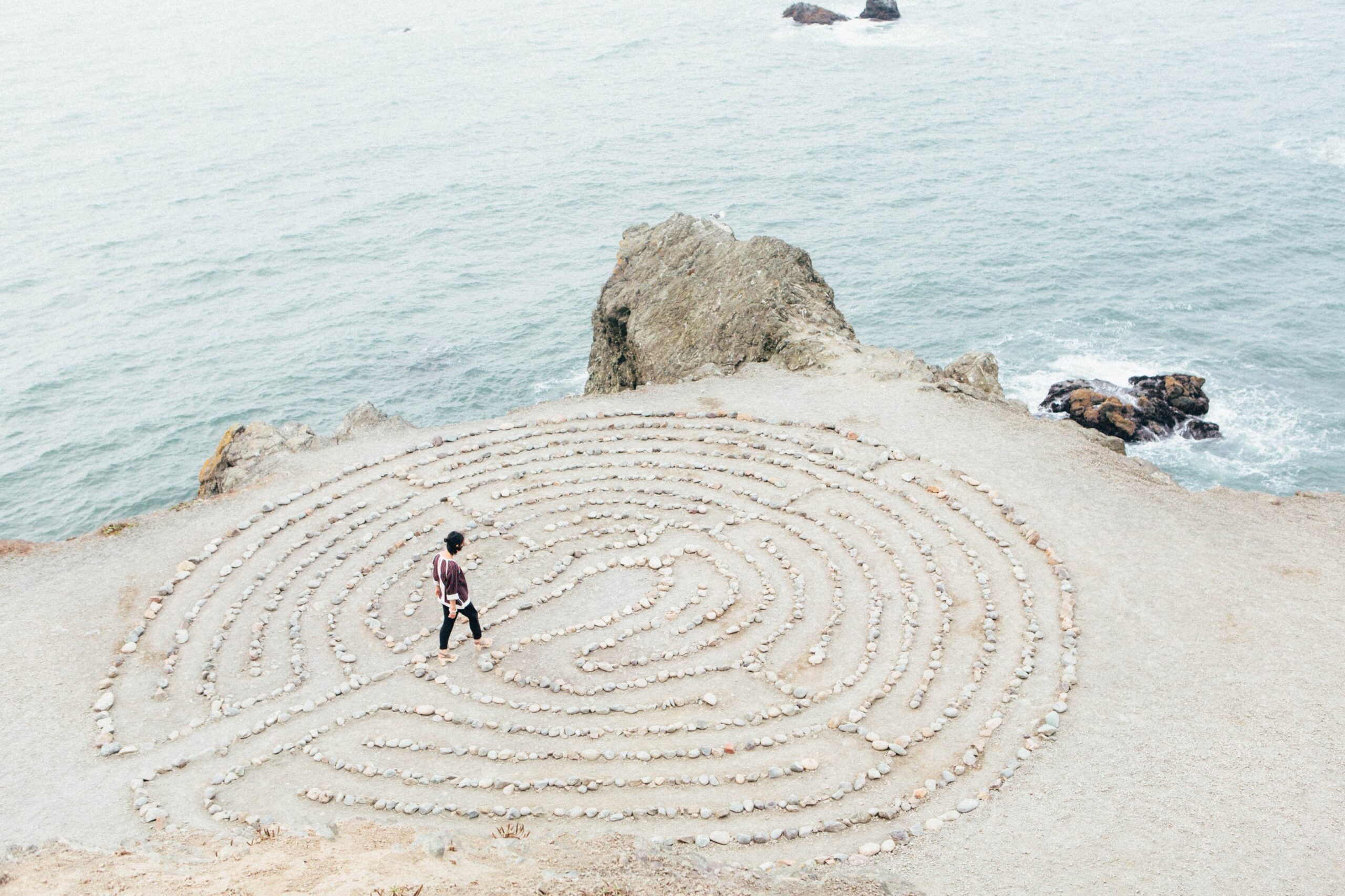 A person walking through a small stone maze on a sandy cliff near the water