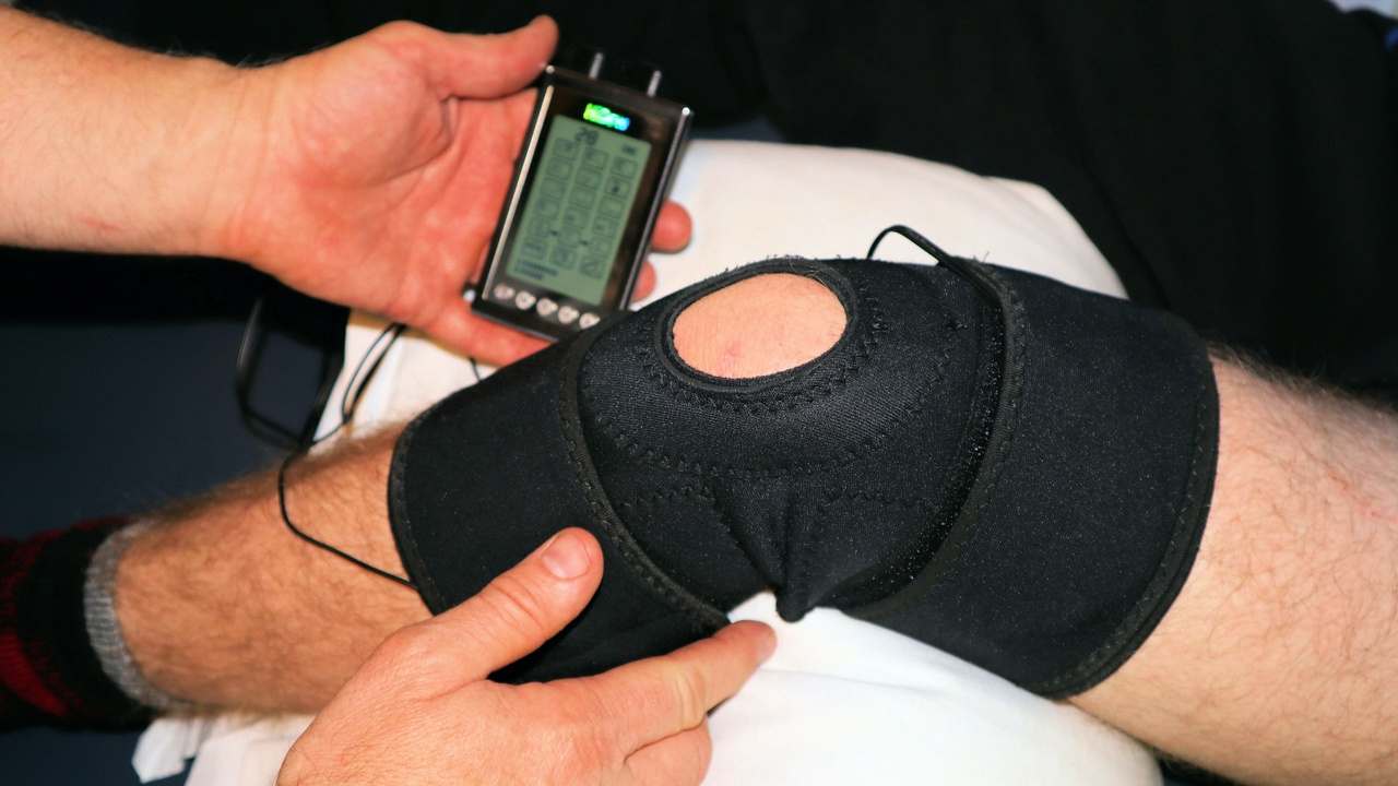 A leg with a black knee brace being assessed by two hands using a electronic device