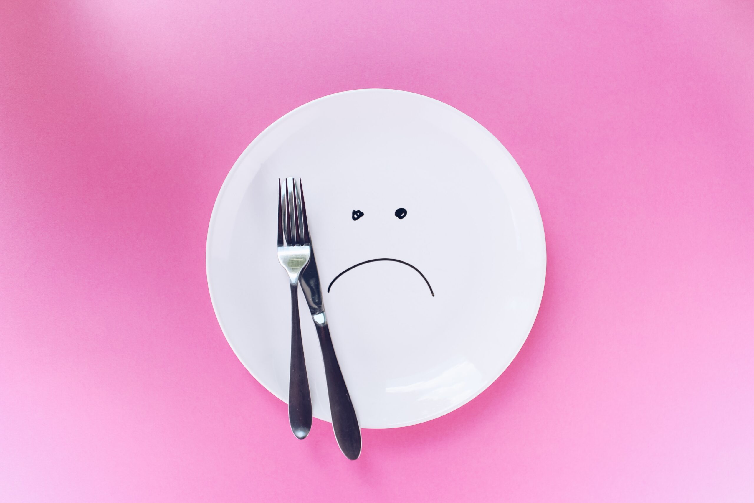 A white dinner plate with a sad face drawn on it, with a silver knife and fork on a pink background