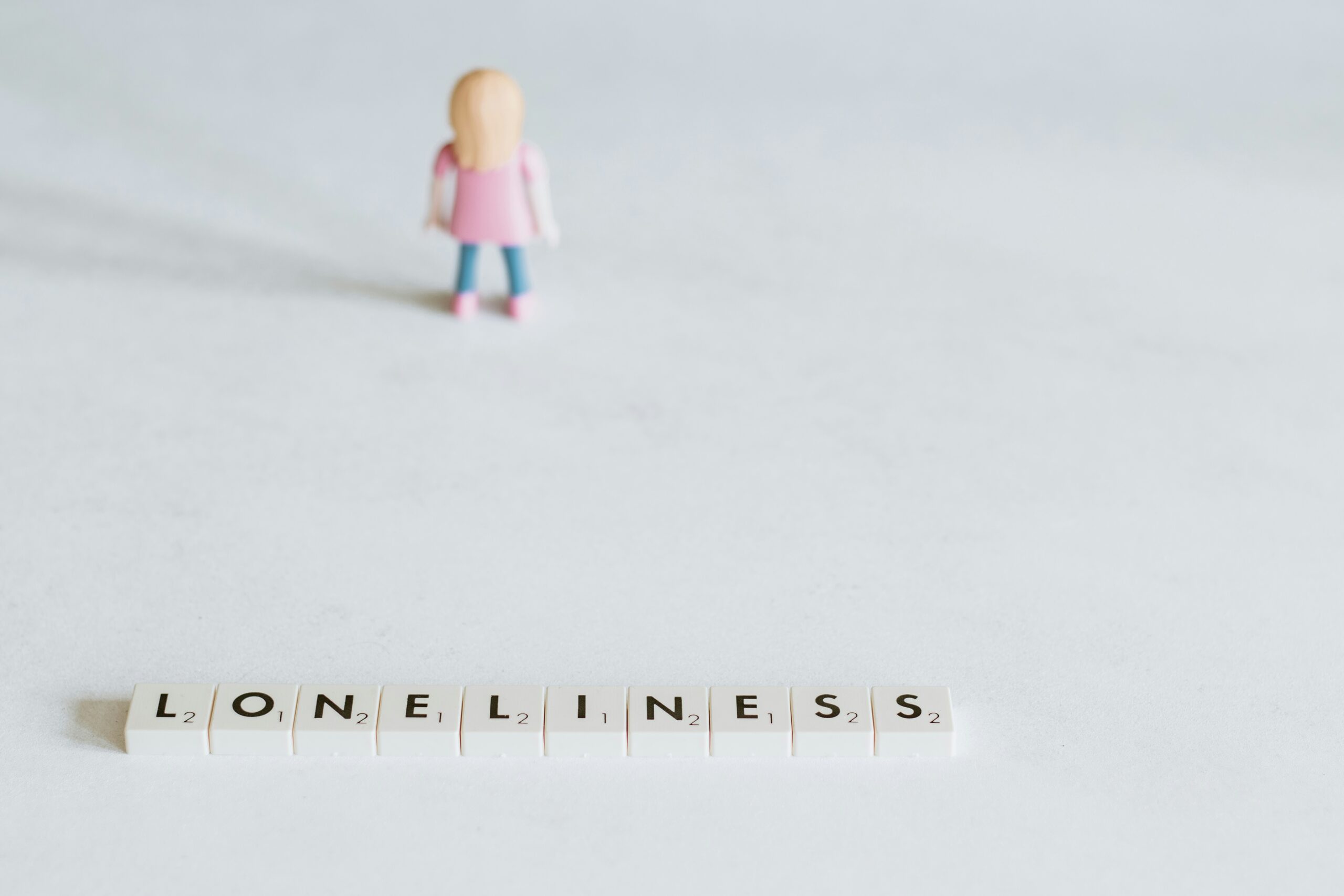 Cream scrabble tiles that spell 'loneliness' laying on a white background. There is a small girl toy facing backwards in the background.