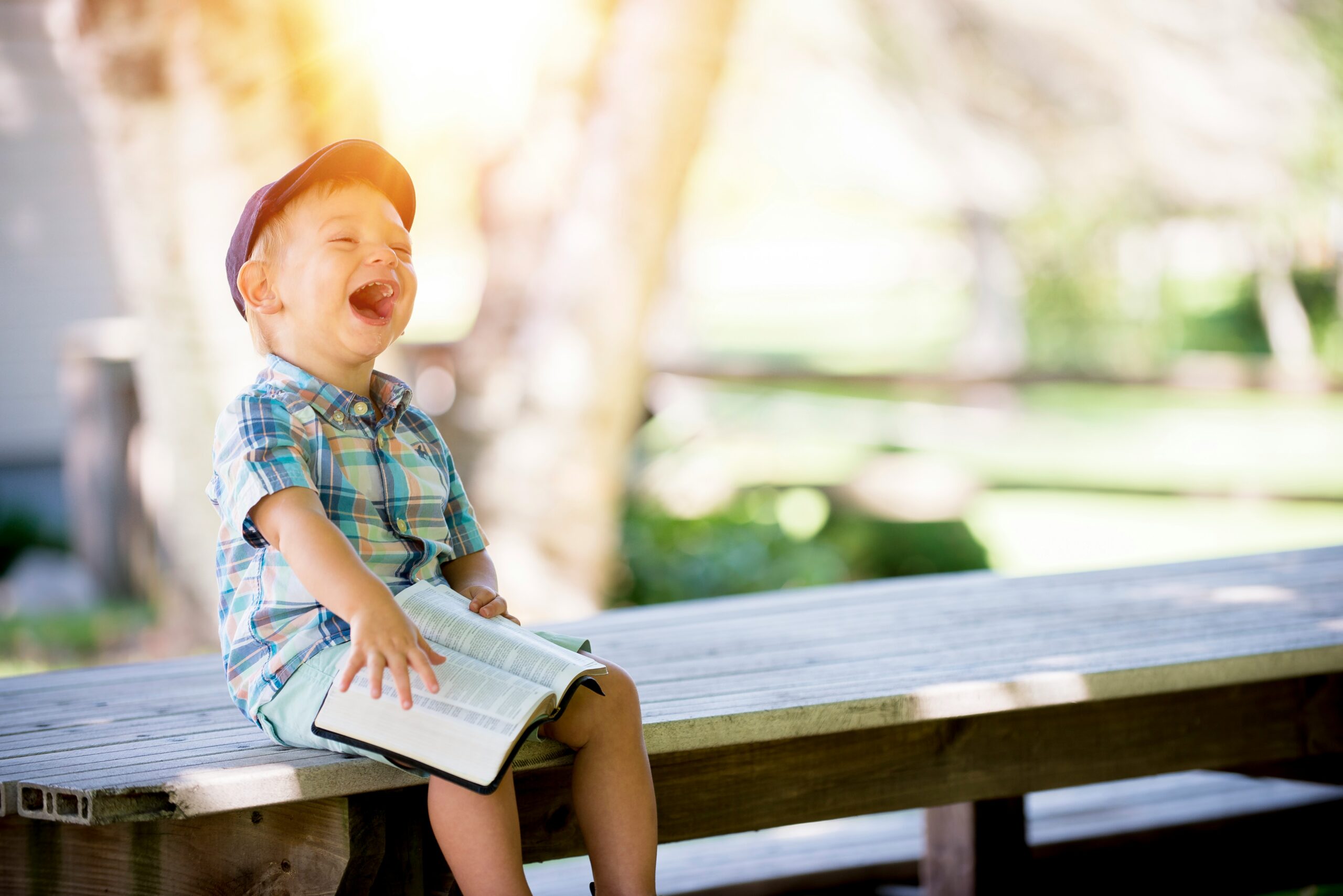 A young boy sitting outside on a wooden bench, laughing with an open book on his lap