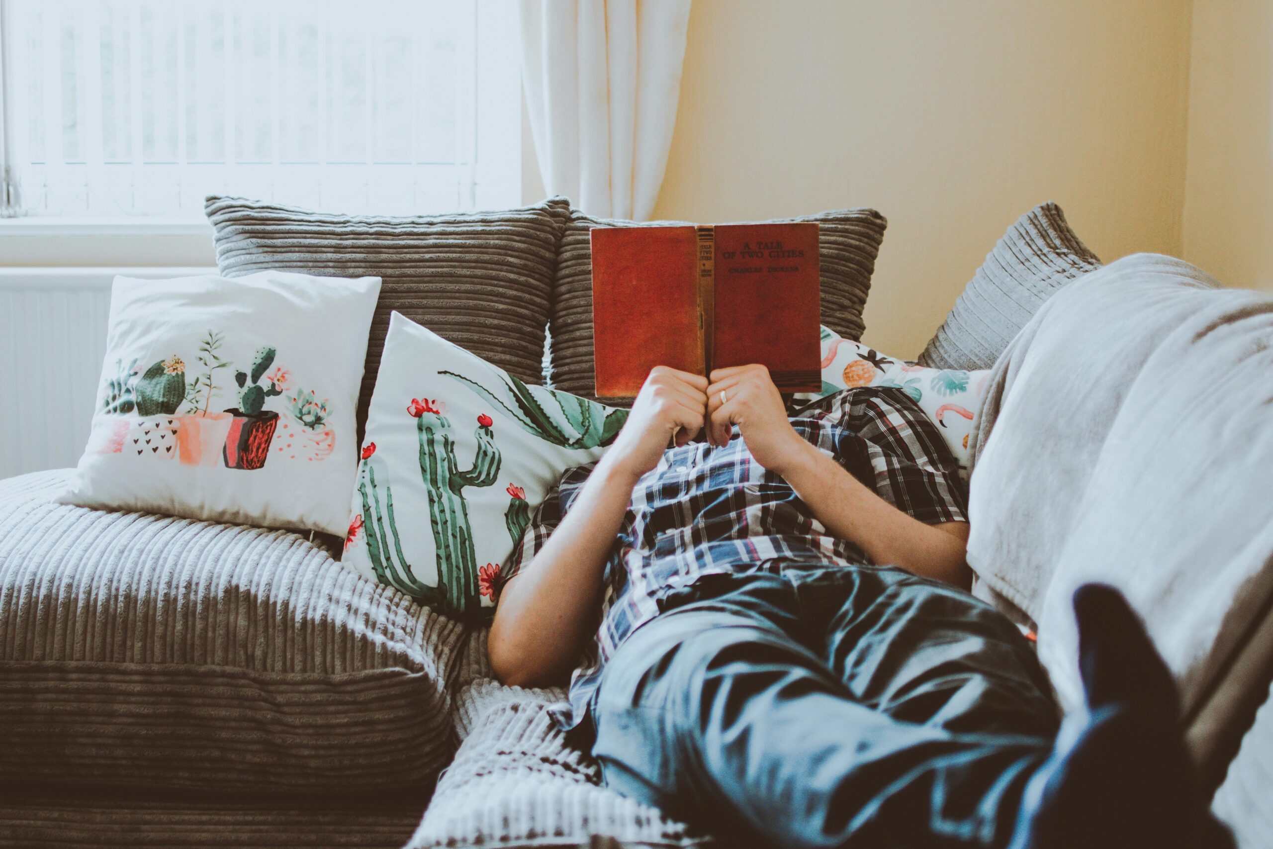 A person wearing denim jeans and a check shirt, lying on a grey couch holding a red book in front of their face.