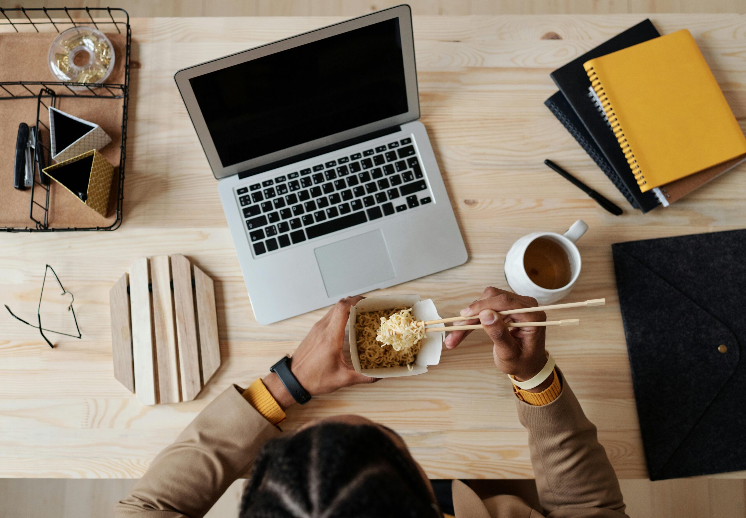 An aerial view of a timber desk with a silver laptop, white mug of coffee and a mans arms and hands using chopsticks to eat noodles out of a white container